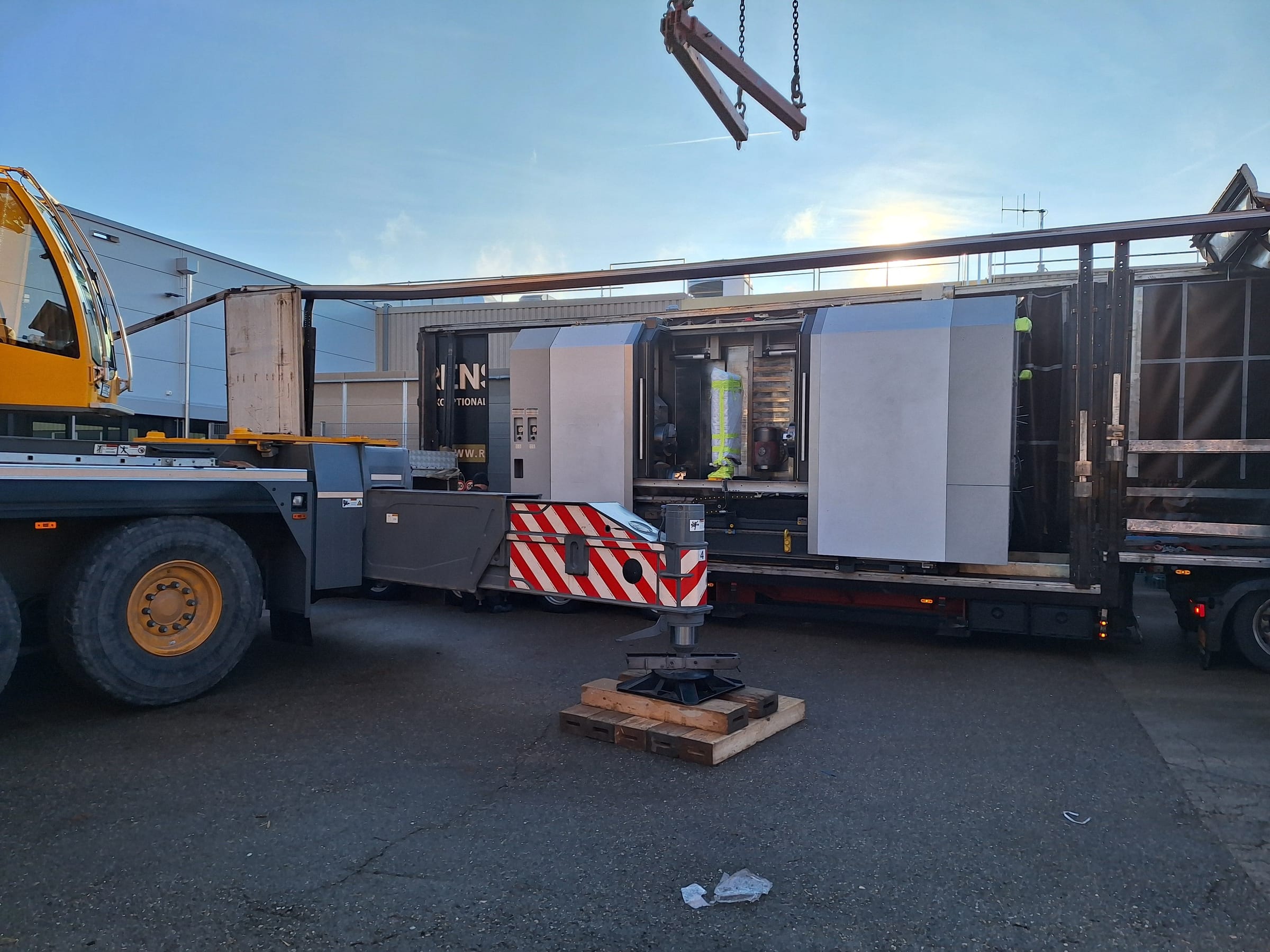 Grúa móvil con estabilizadores junto a un semirremolque con una máquina herramienta cargada